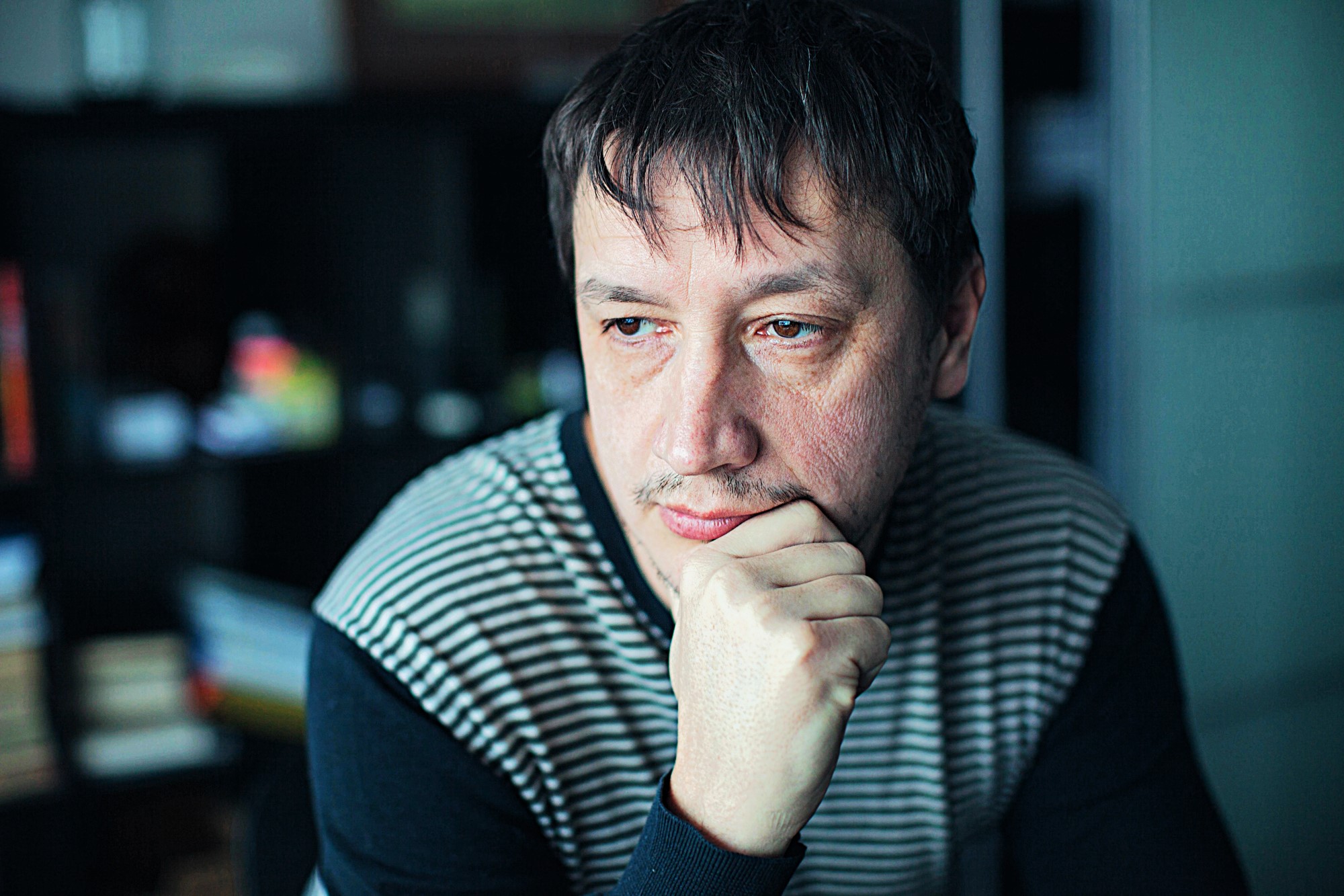 A man with dark hair and facial hair, wearing a striped sweater, is seated indoors. He rests his chin on his hand, appearing thoughtful, with a blurred background featuring shelves and books.