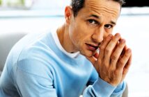 A man in a light blue sweater rests his chin on his hands, looking intently at the camera with a thoughtful expression. The background is blurred, emphasizing the man’s face and contemplative pose.