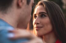 A woman with long brown hair gazes intently at a man, her face showing a soft smile. The man's face is blurred and turned slightly to the side. They appear to be outdoors, with a soft, warm background.