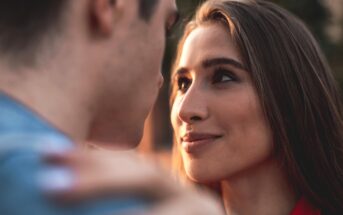 A woman with long brown hair gazes intently at a man, her face showing a soft smile. The man's face is blurred and turned slightly to the side. They appear to be outdoors, with a soft, warm background.