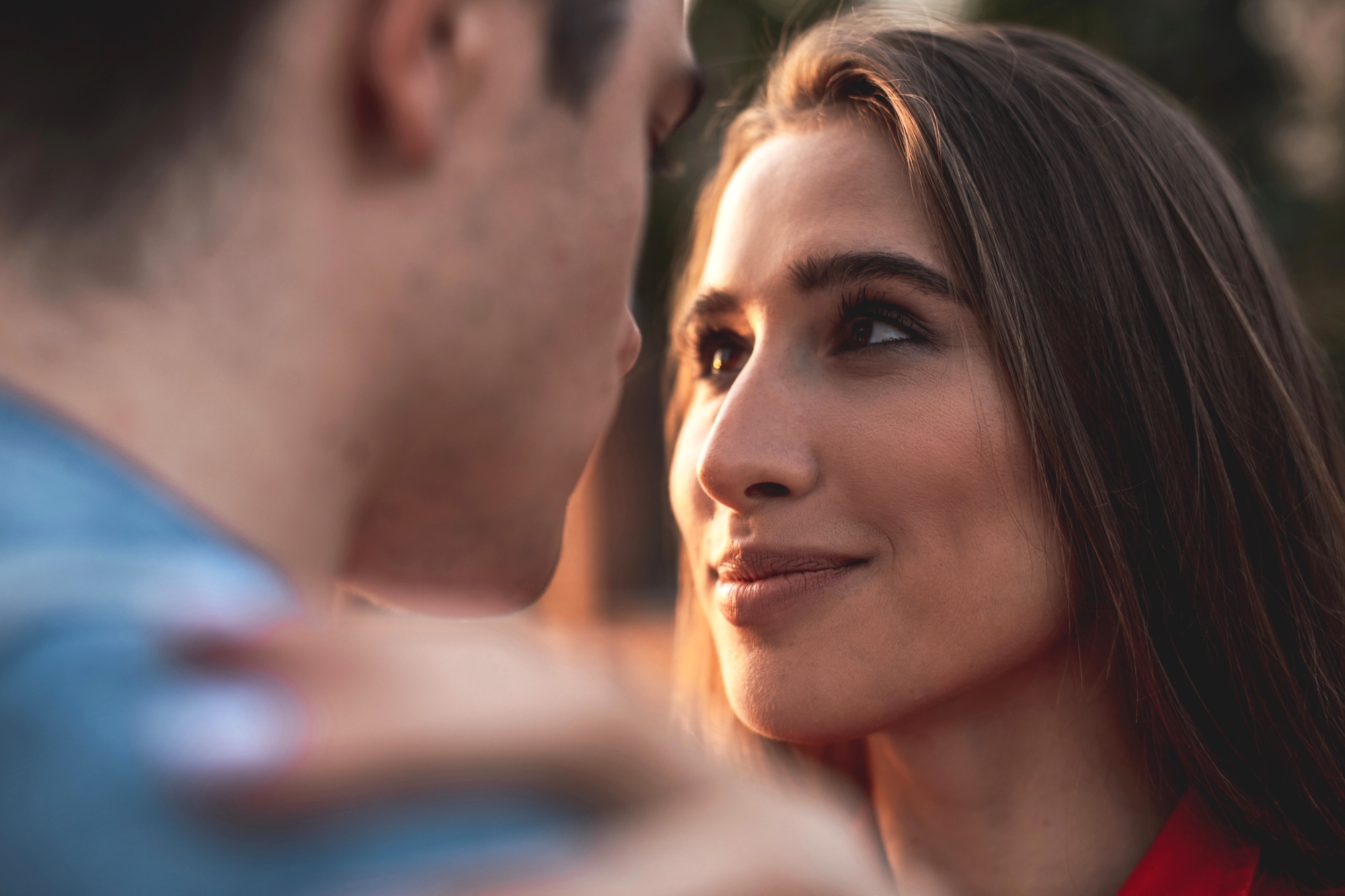 A woman with long brown hair gazes intently at a man, her face showing a soft smile. The man's face is blurred and turned slightly to the side. They appear to be outdoors, with a soft, warm background.