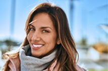 A person with long brown hair smiles warmly, wearing a beige coat and gray scarf. The background shows a blurred outdoor scene with bright sunlight and blue sky.