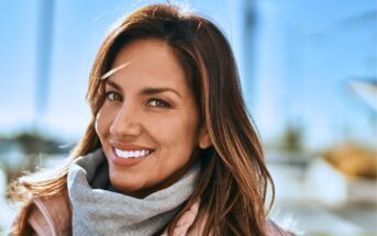 A person with long brown hair smiles warmly, wearing a beige coat and gray scarf. The background shows a blurred outdoor scene with bright sunlight and blue sky.