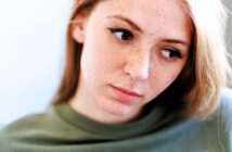 A woman with light skin and freckles looks pensively to the side. She has long brown hair and is wearing a green top. The background is softly blurred, emphasizing her thoughtful expression.