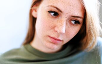 A woman with light skin and freckles looks pensively to the side. She has long brown hair and is wearing a green top. The background is softly blurred, emphasizing her thoughtful expression.