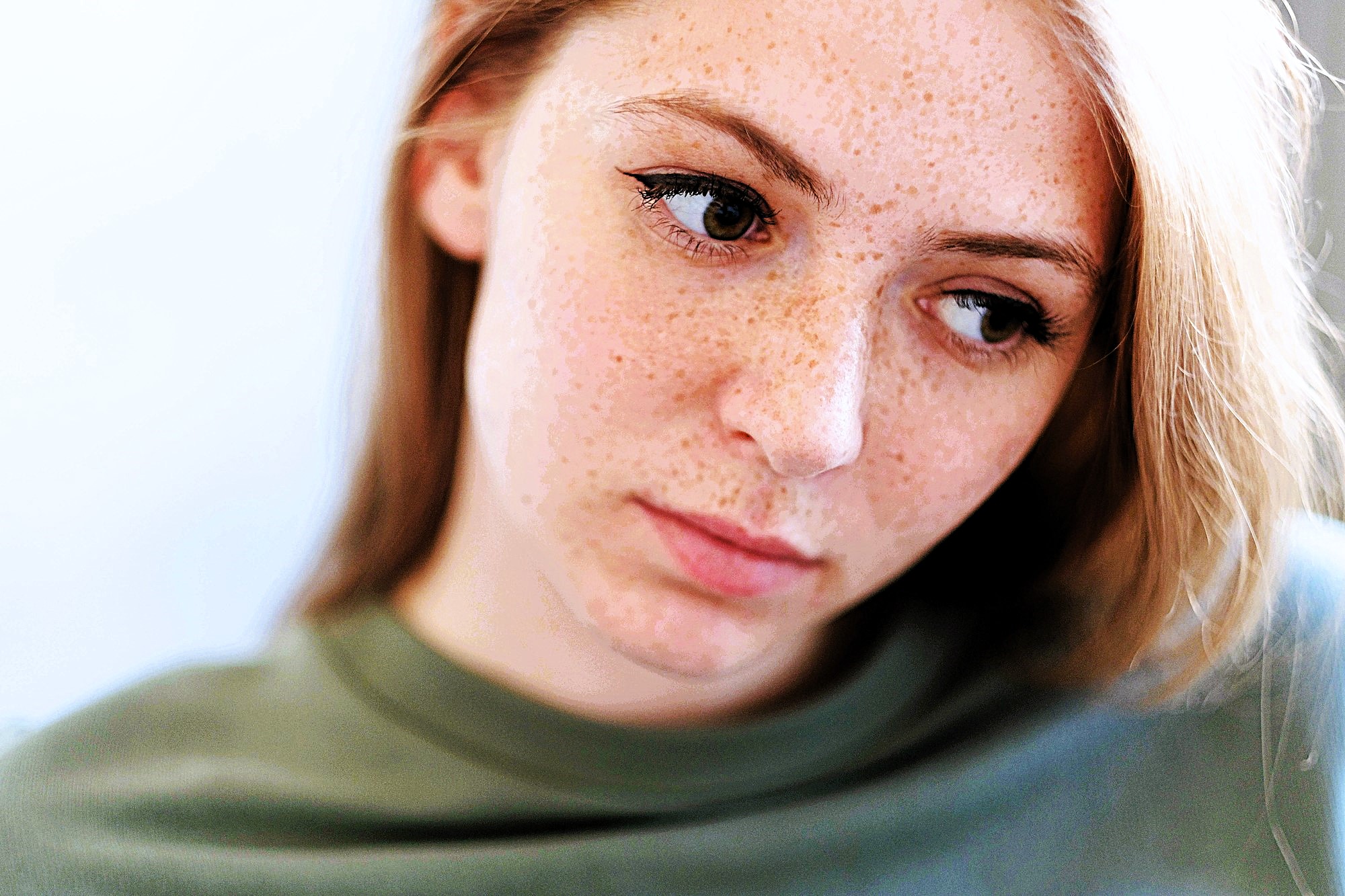 A woman with light skin and freckles looks pensively to the side. She has long brown hair and is wearing a green top. The background is softly blurred, emphasizing her thoughtful expression.