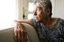 Elderly woman with short gray hair wearing a patterned gray shirt, sitting on a couch, gazing thoughtfully out of a sunlit window. Her hand rests on the couch back, and she appears deep in thought.