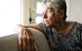 Elderly woman with short gray hair wearing a patterned gray shirt, sitting on a couch, gazing thoughtfully out of a sunlit window. Her hand rests on the couch back, and she appears deep in thought.