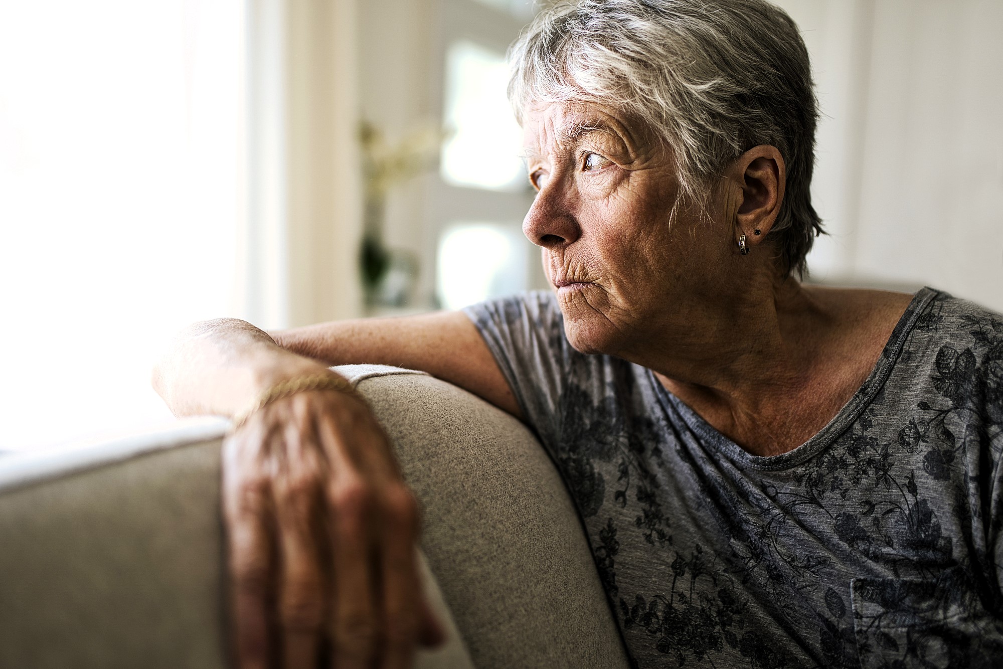 Elderly woman with short gray hair wearing a patterned gray shirt, sitting on a couch, gazing thoughtfully out of a sunlit window. Her hand rests on the couch back, and she appears deep in thought.