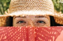 A person with blue eyes and freckles is looking over an open red book, wearing a straw hat that casts a shadow pattern on their face. The background is out of focus, suggesting a sunny outdoor setting.