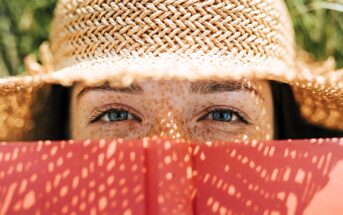 A person with blue eyes and freckles is looking over an open red book, wearing a straw hat that casts a shadow pattern on their face. The background is out of focus, suggesting a sunny outdoor setting.