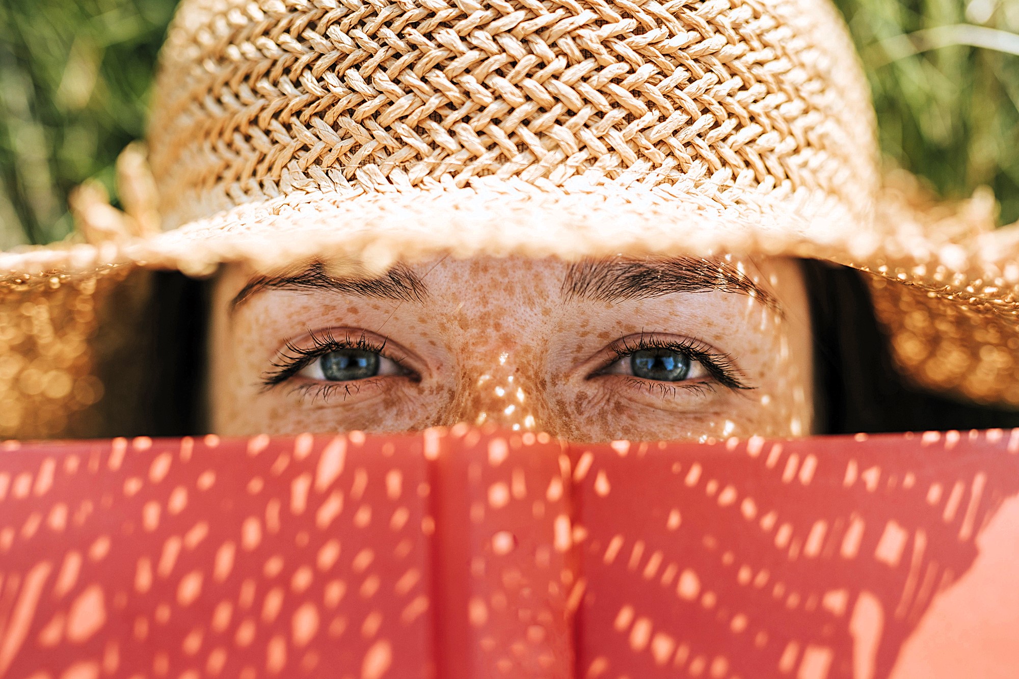 A person with blue eyes and freckles is looking over an open red book, wearing a straw hat that casts a shadow pattern on their face. The background is out of focus, suggesting a sunny outdoor setting.