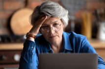 An older adult with short gray hair and glasses looks thoughtfully at a laptop screen. They are wearing a blue shirt and resting their head on one hand, seated in a cozy kitchen setting.