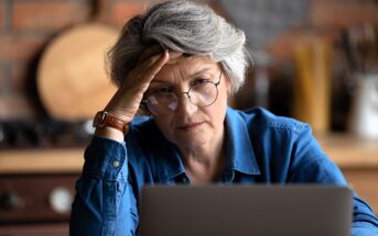 An older adult with short gray hair and glasses looks thoughtfully at a laptop screen. They are wearing a blue shirt and resting their head on one hand, seated in a cozy kitchen setting.