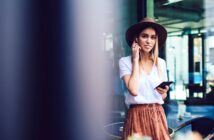 A woman wearing a hat, a white shirt, and a brown skirt is standing outside, holding a smartphone and listening to music with earbuds. She is smiling and appears relaxed, with a cityscape in the background.