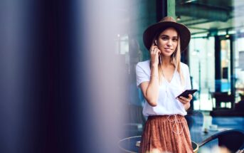 A woman wearing a hat, a white shirt, and a brown skirt is standing outside, holding a smartphone and listening to music with earbuds. She is smiling and appears relaxed, with a cityscape in the background.