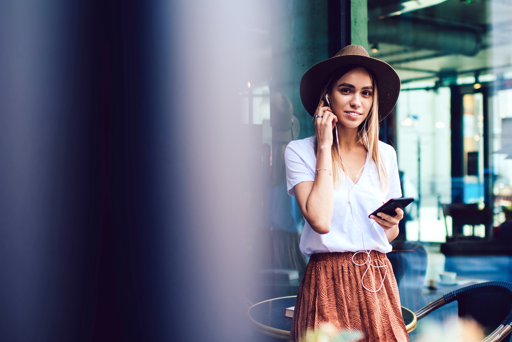 A woman wearing a hat, a white shirt, and a brown skirt is standing outside, holding a smartphone and listening to music with earbuds. She is smiling and appears relaxed, with a cityscape in the background.