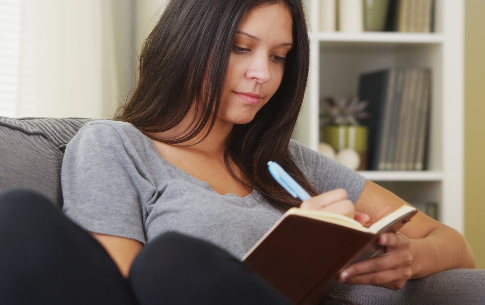 A woman with long dark hair sits on a couch, writing in a brown notebook with a blue pen. She is wearing a gray T-shirt and appears focused. In the background, there is a white bookshelf with various books and a small plant.