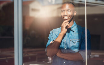 A person in a denim shirt, standing indoors with a reflective window. They are smiling and resting their chin on their hand, looking thoughtful. Sunlight is streaming in, creating a warm ambiance.