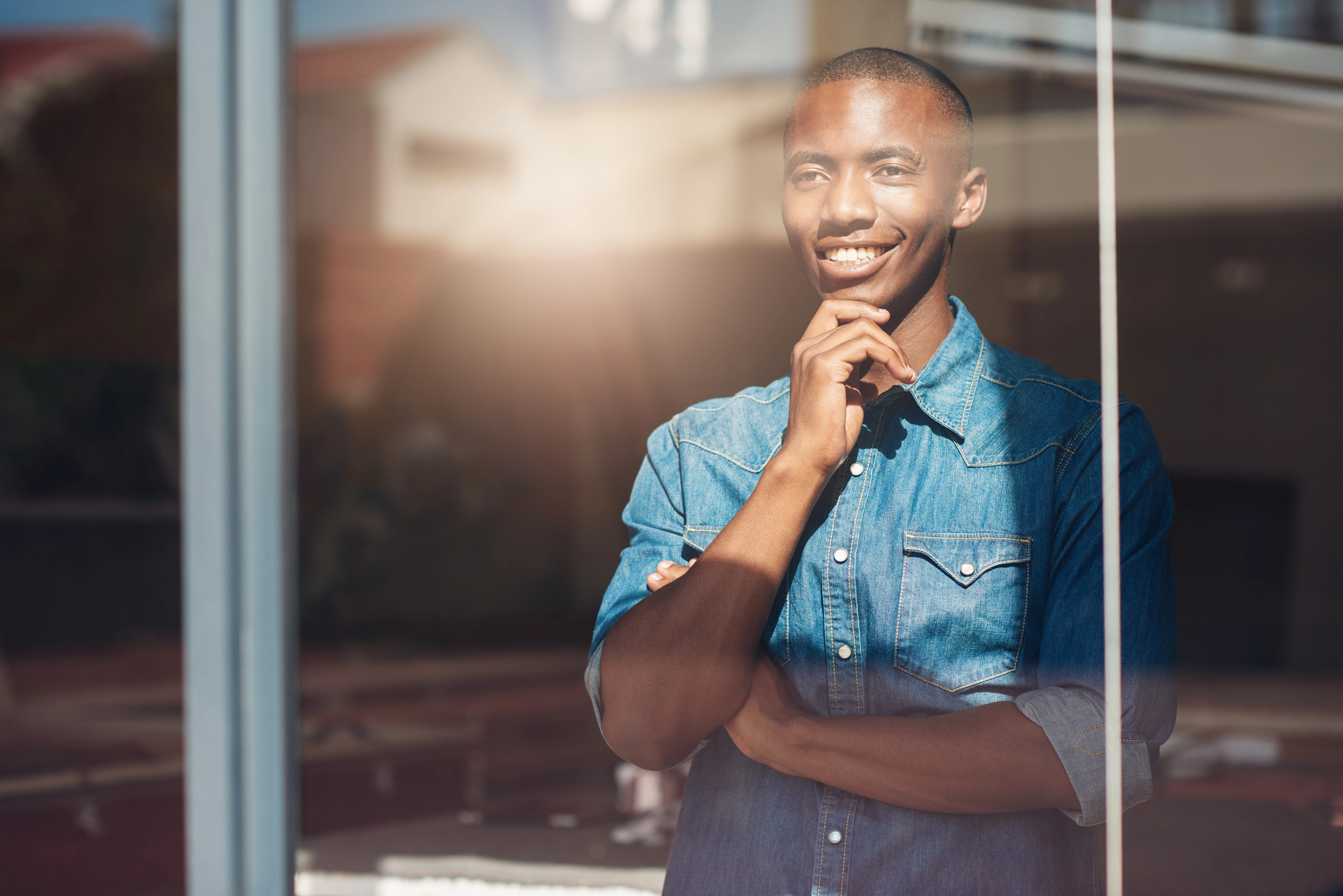 A person in a denim shirt, standing indoors with a reflective window. They are smiling and resting their chin on their hand, looking thoughtful. Sunlight is streaming in, creating a warm ambiance.