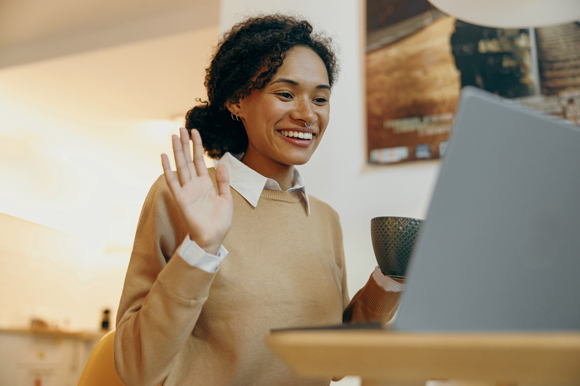 A person sits at a table, smiling and waving at a laptop screen. They hold a mug in their other hand. The background is a cozy home setting with warm lighting and a blurred poster on the wall.