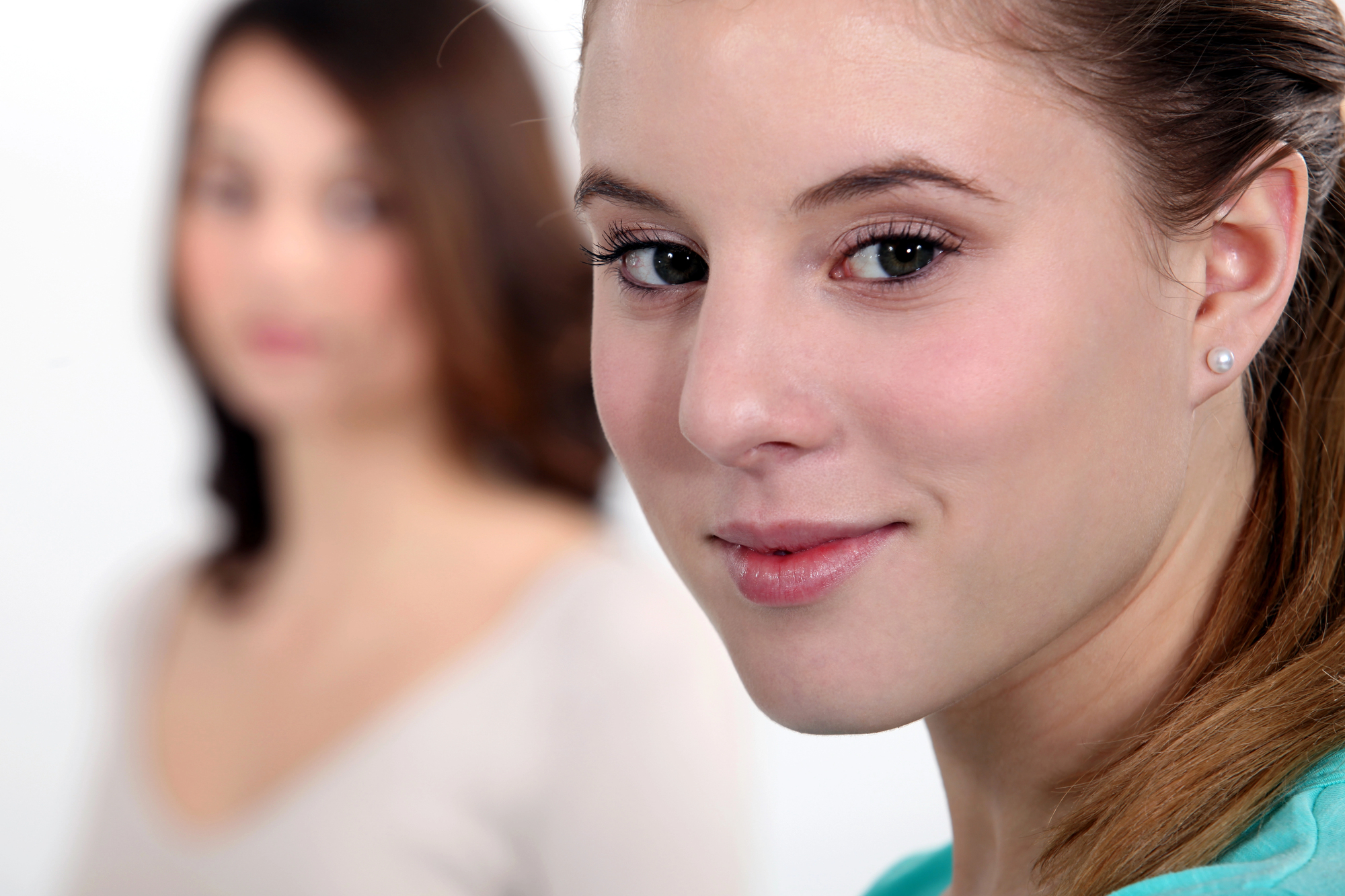 Close-up of a smiling young woman in focus, with another woman in the background out of focus. The young woman has light brown hair tied back and is wearing a light blue top. The background is plain and white.