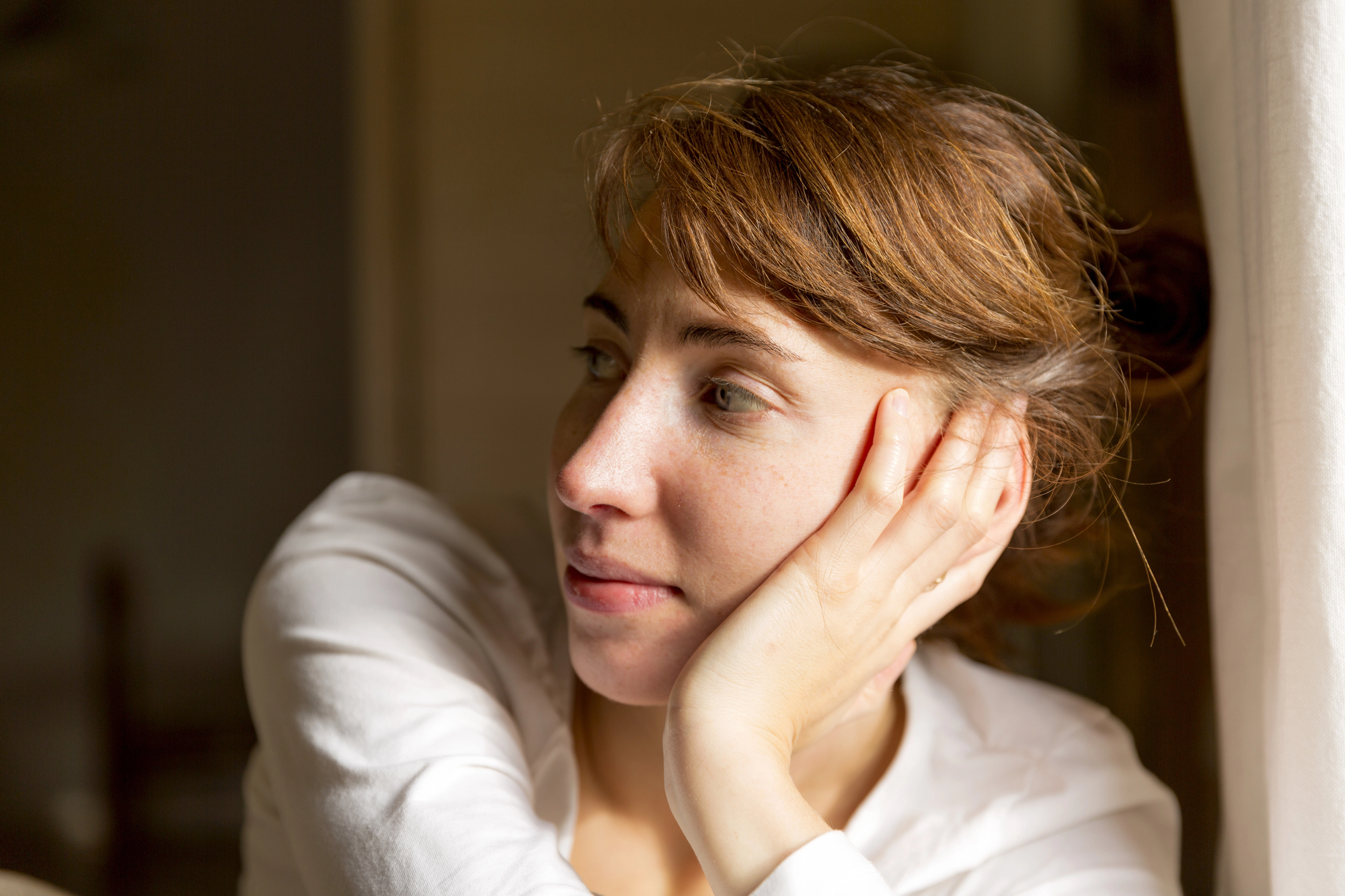 A woman with brown hair, wearing a white shirt, rests her chin on her hand while gazing thoughtfully out of a window. Soft sunlight illuminates her face, creating a warm and serene atmosphere.