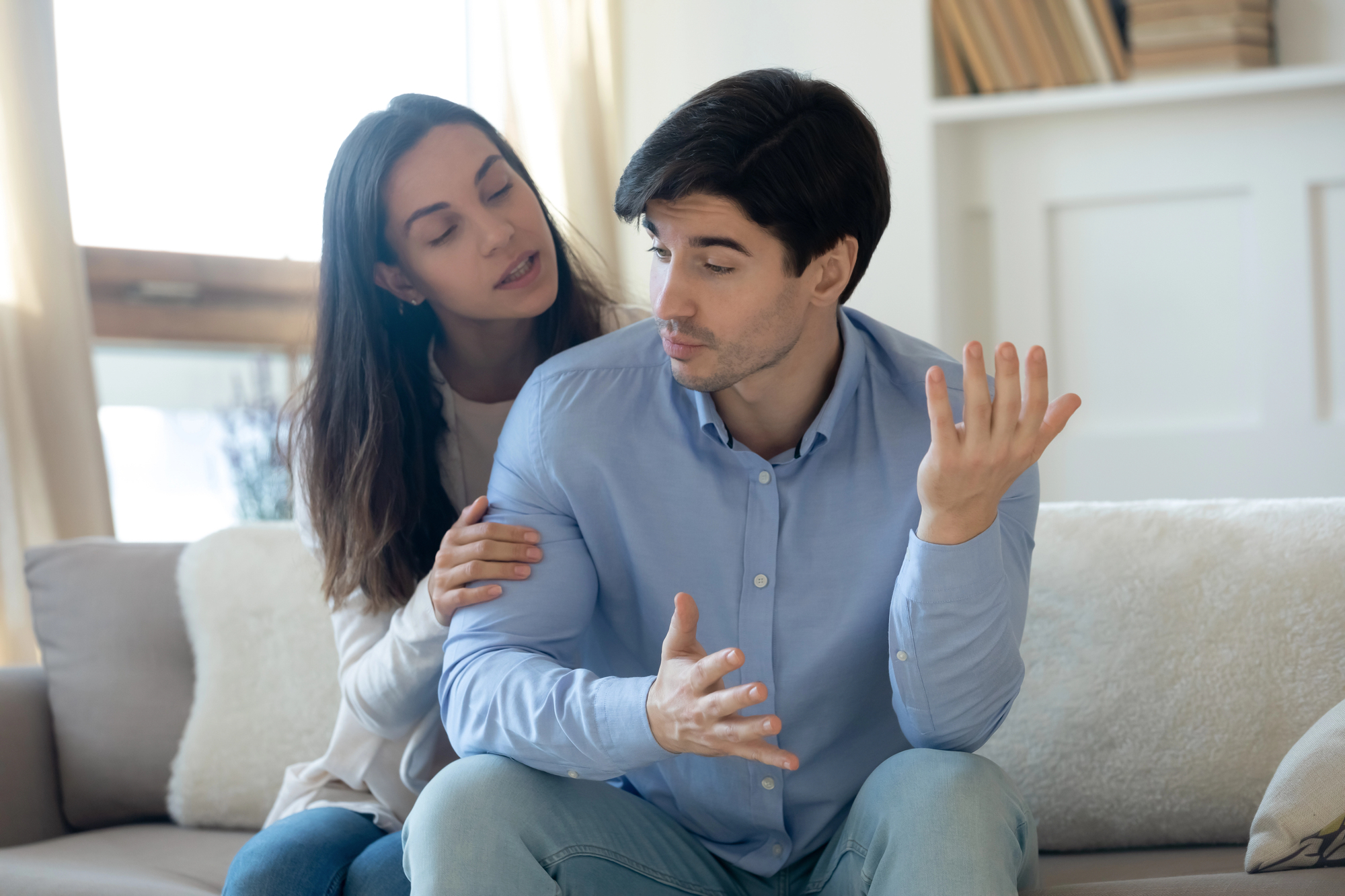 A woman is sitting on a couch, leaning towards a man who appears to be explaining or discussing something with hand gestures. They are indoors, with a window and bookshelf in the background. The mood seems engaged and conversational.