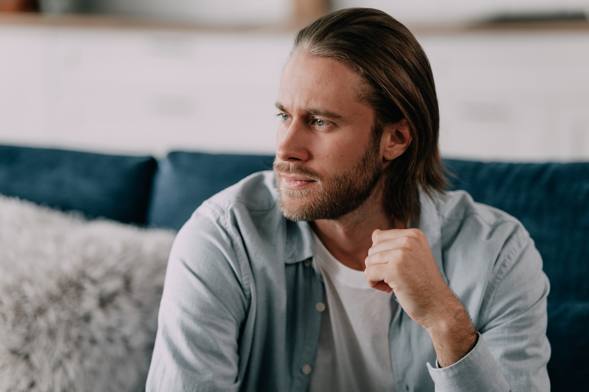 A man with long hair and a beard sits on a blue couch, looking thoughtfully into the distance. He is wearing a light blue shirt over a white T-shirt. A fluffy gray pillow is visible next to him. The background is a blurred white room.