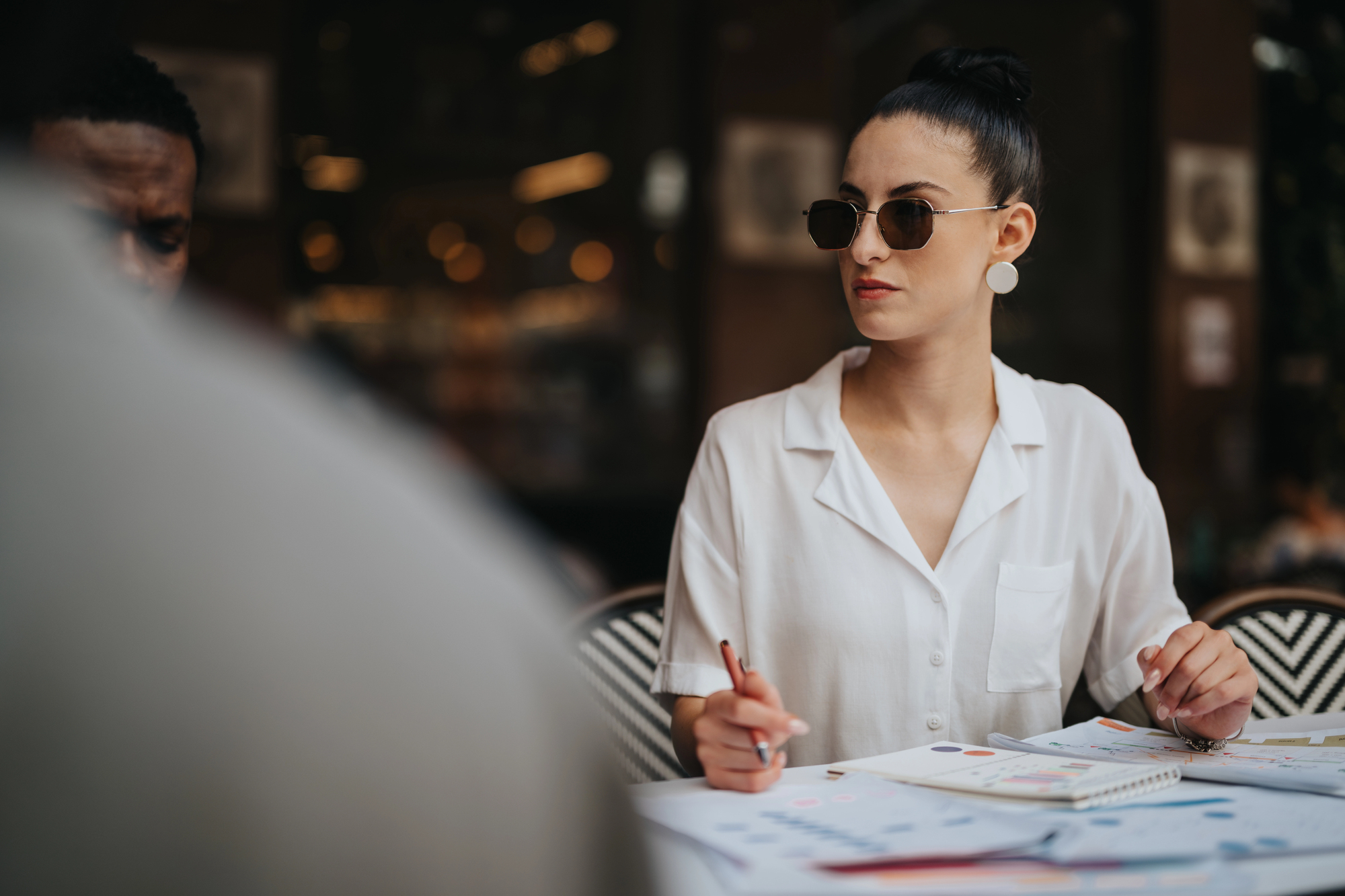 A woman wearing sunglasses and a white blouse sits at a table with documents and colorful charts. She is holding a pen and looking attentively to her side. The background is dimly lit, with blurred lights.