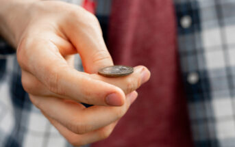 A person holding a coin between their thumb and index finger, wearing a checkered shirt and a red top underneath. The focus is on the hand and the coin.