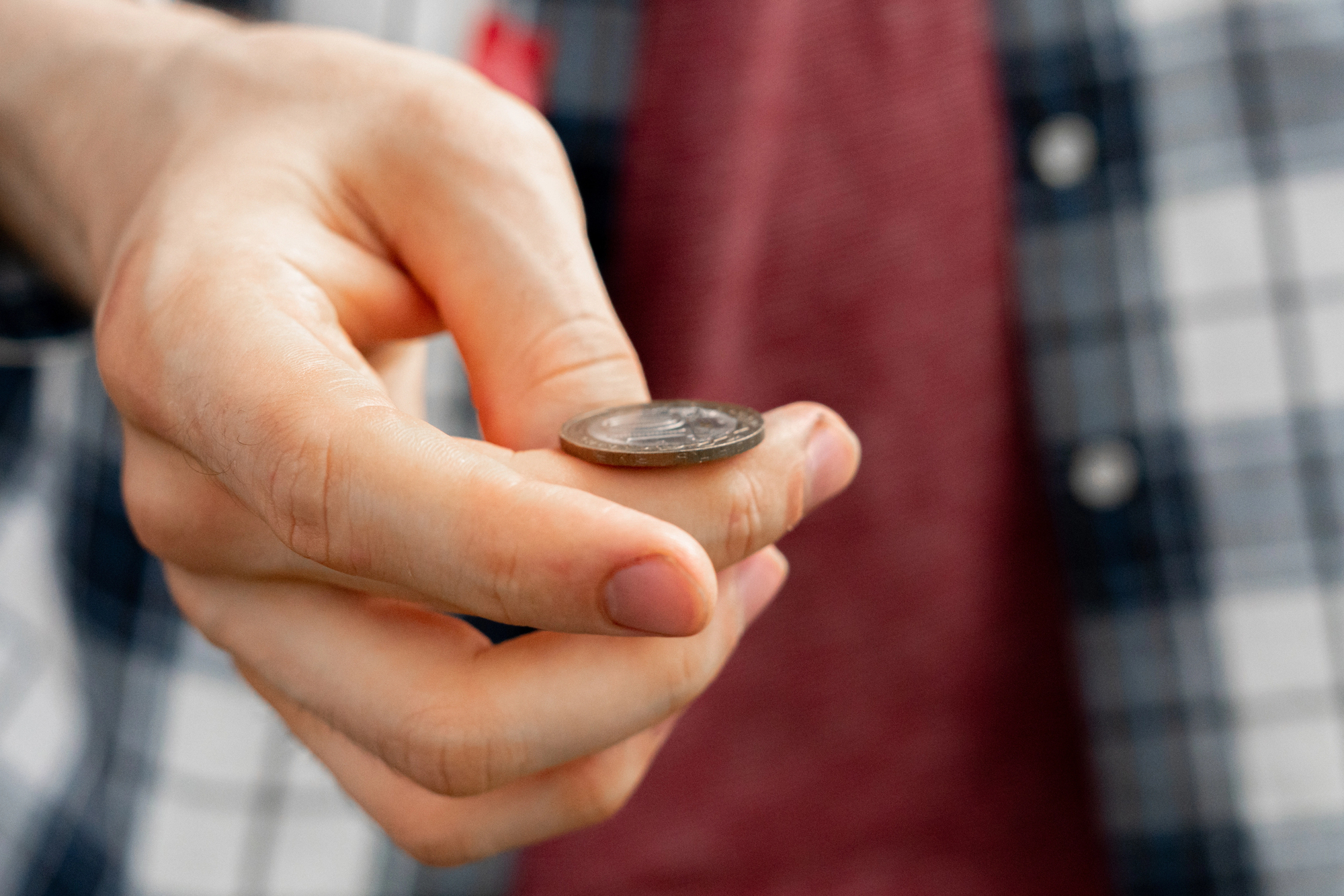 A person holding a coin between their thumb and index finger, wearing a checkered shirt and a red top underneath. The focus is on the hand and the coin.