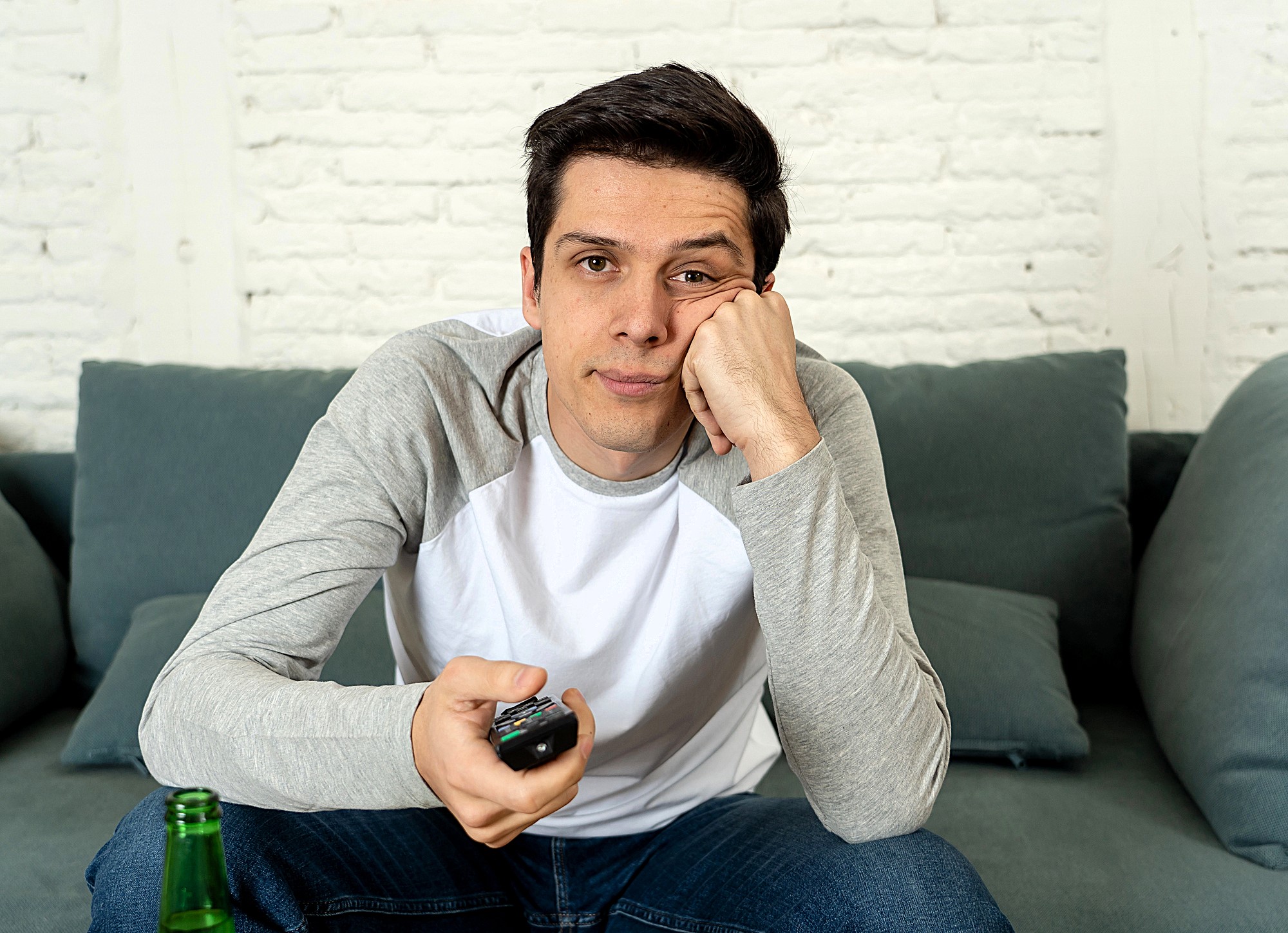 A man sits on a couch holding a remote control, resting his chin on his hand, looking bored. He wears a gray and white long-sleeve shirt, with a green bottle on the table in front of him, and a white brick wall in the background.