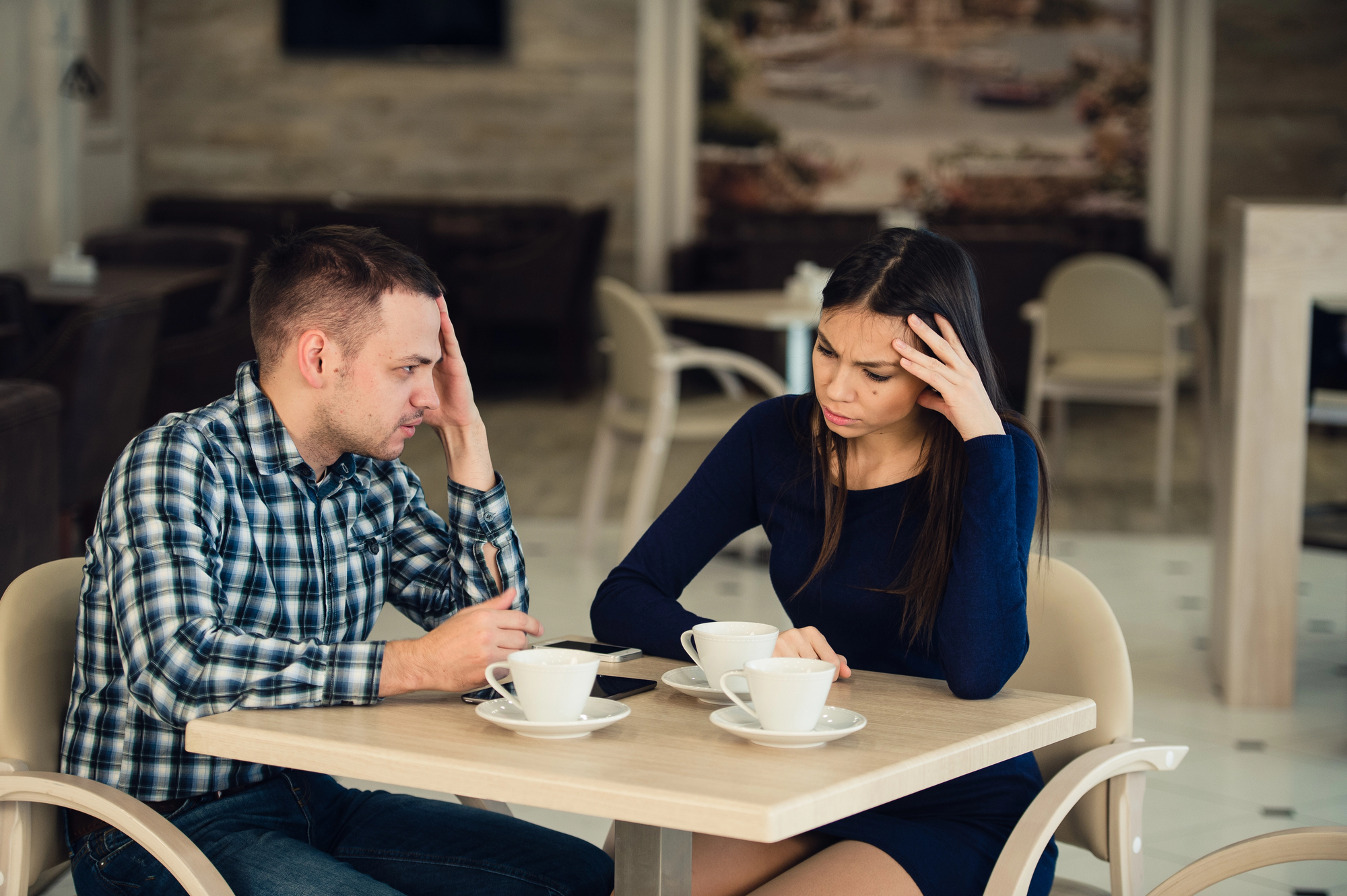 A man and woman sit at a café table, both looking stressed as they hold their heads with one hand. Two cups of coffee and a smartphone are on the table. The café interior is softly lit and decorated in neutral colors.