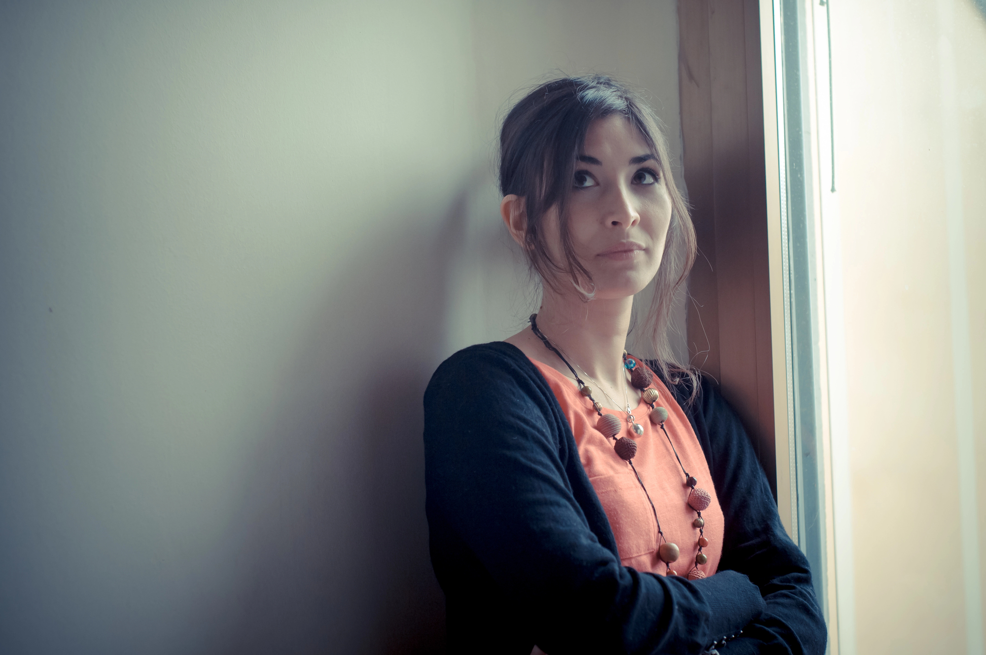 A woman with long hair stands by a window, wearing a coral top and black cardigan. She has a thoughtful expression, with arms crossed, and wears a beaded necklace. Soft light illuminates her face against a neutral wall.