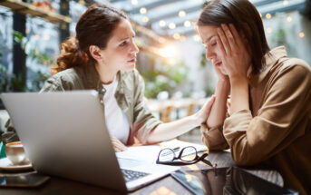 A woman comforts a distressed friend at a table in a cafe. They are sitting in front of a laptop, with eyeglasses and papers scattered nearby. The setting is bright with blurred lights and greenery in the background.