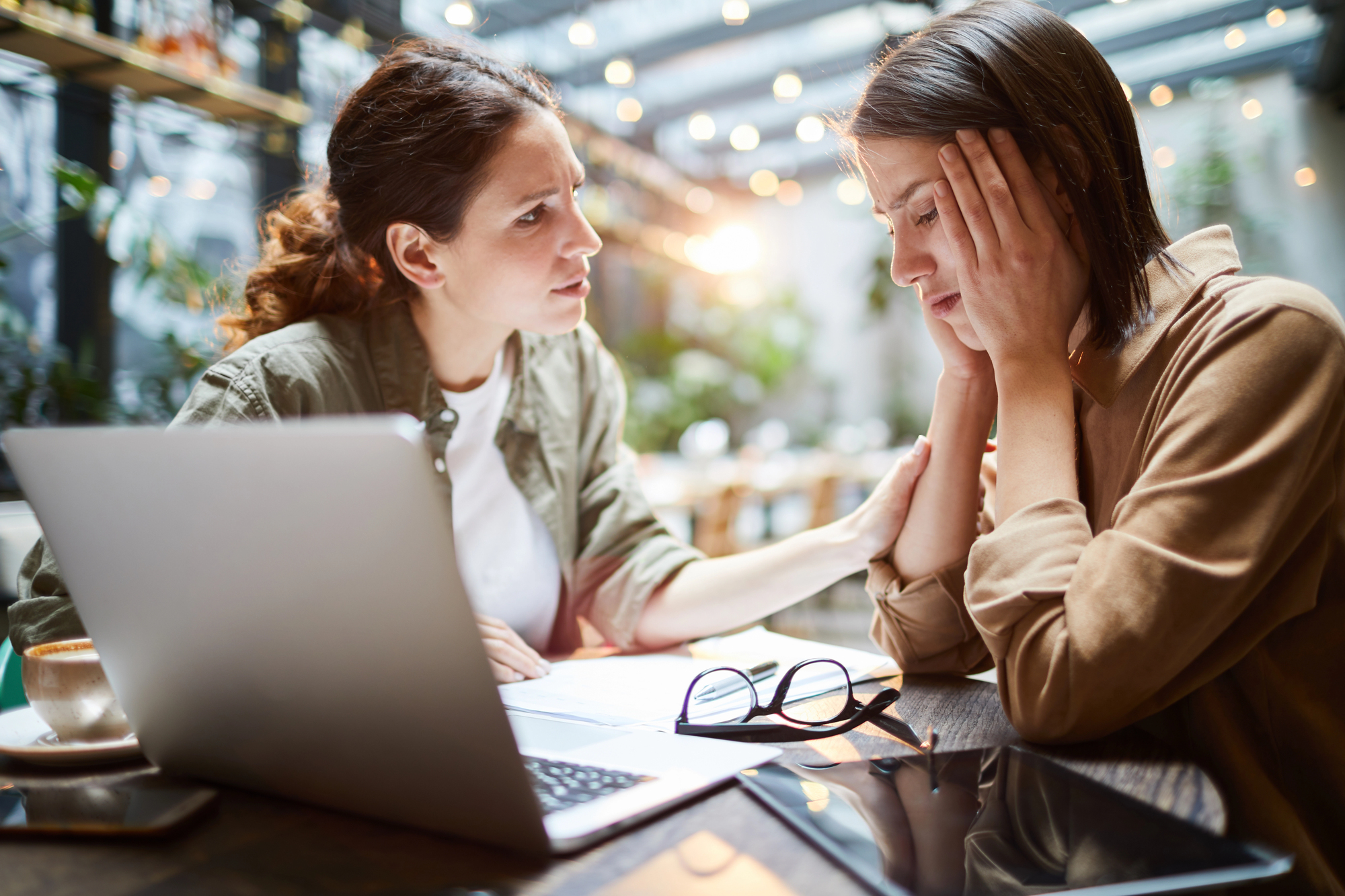 A woman comforts a distressed friend at a table in a cafe. They are sitting in front of a laptop, with eyeglasses and papers scattered nearby. The setting is bright with blurred lights and greenery in the background.