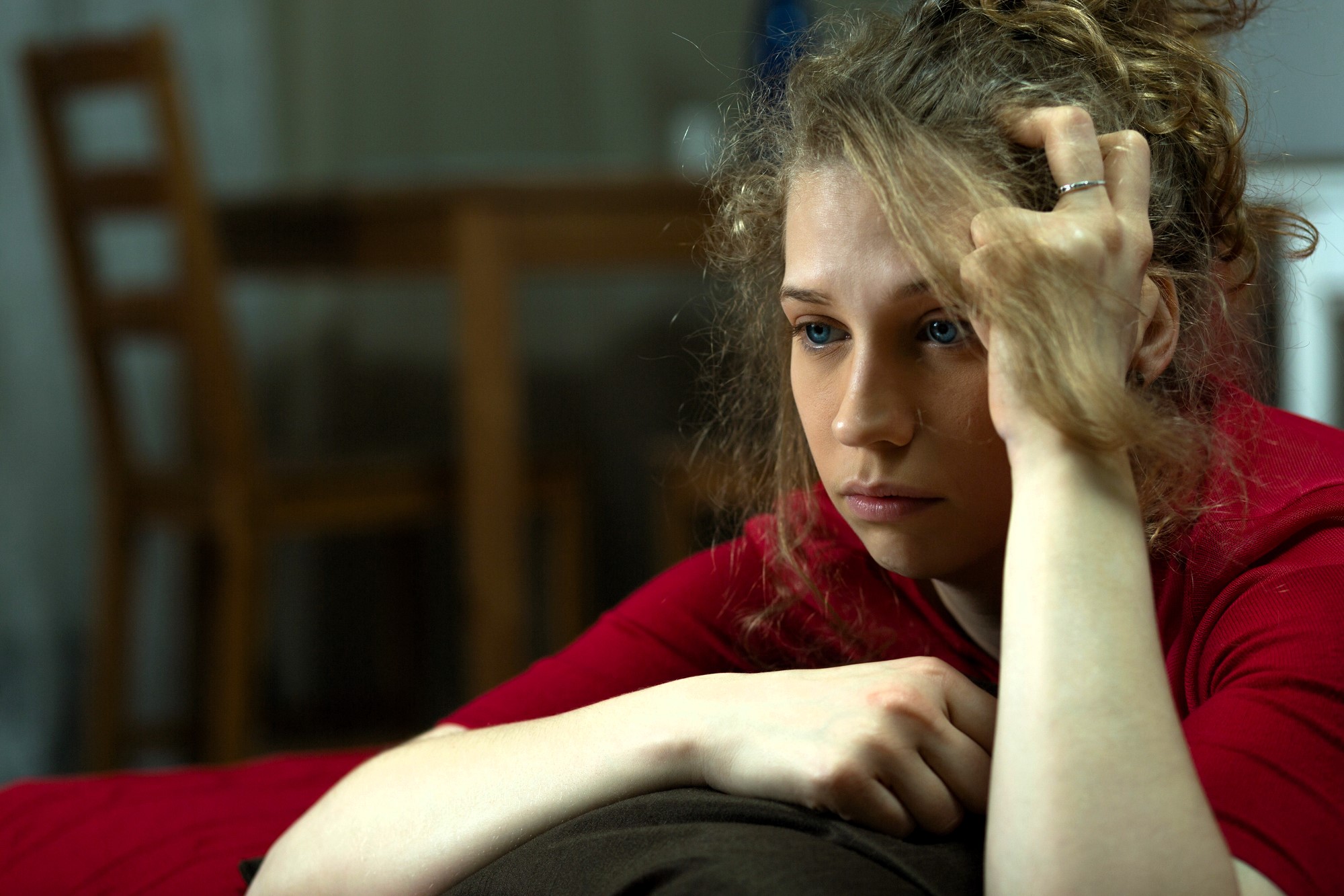 A woman with curly hair wearing a red shirt sits with her head resting on her hand, looking pensive. A wooden chair and table are blurred in the background.