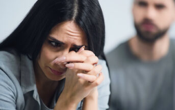 A woman with long dark hair appears distressed, resting her chin on her clasped hands and looking down. A man with a beard is blurred in the background, suggesting tension between them. Both are indoors, wearing casual clothing.