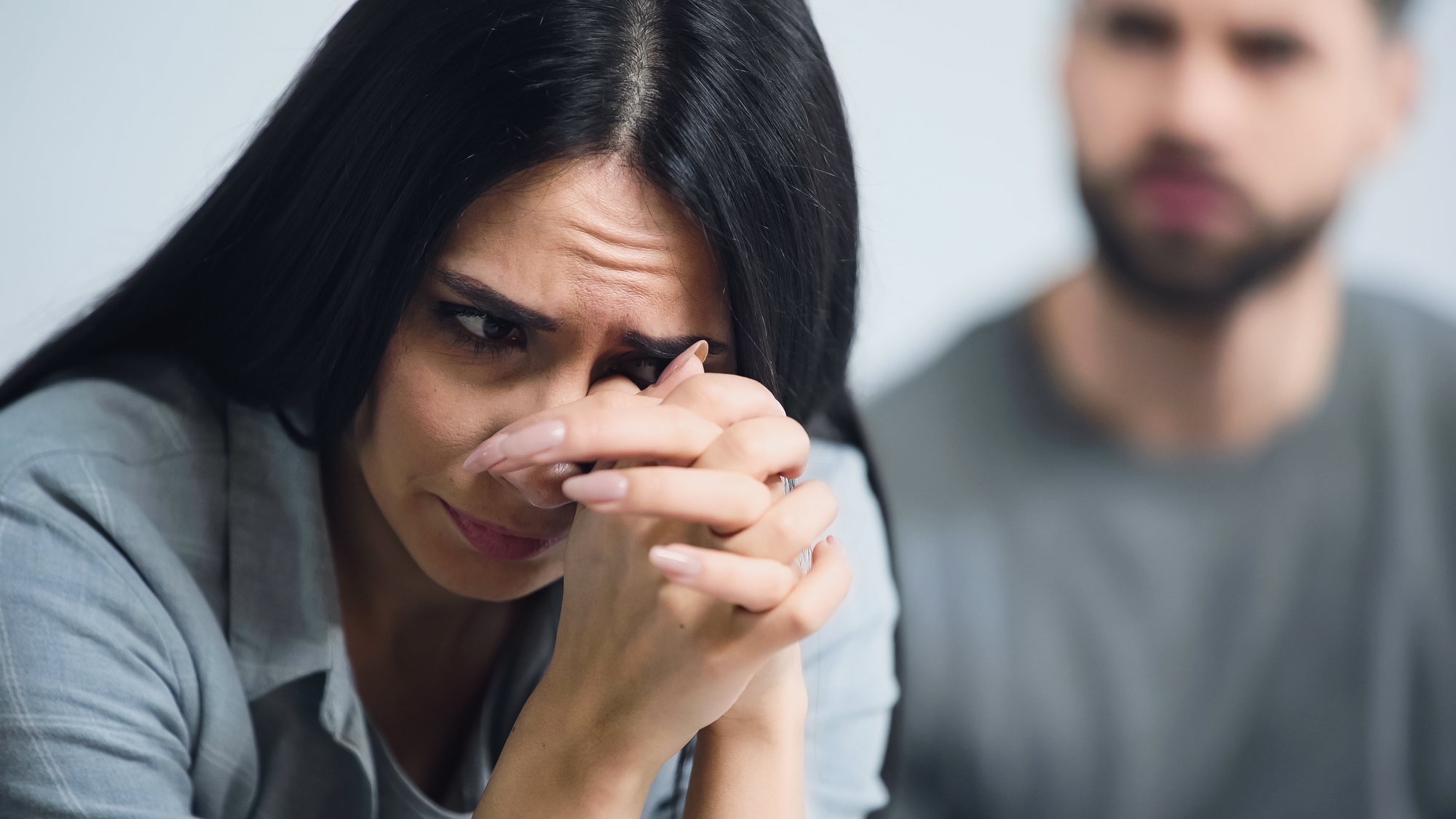 A woman with long dark hair appears distressed, resting her chin on her clasped hands and looking down. A man with a beard is blurred in the background, suggesting tension between them. Both are indoors, wearing casual clothing.