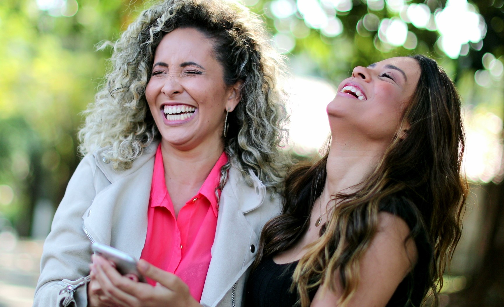 Two women are laughing joyfully outdoors. One has curly hair and wears a light jacket over a pink top, while holding a smartphone. The other has long hair and wears a black top. Bright greenery is blurred in the background.