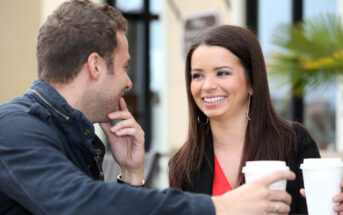 A man and woman are sitting outdoors, smiling and talking while holding coffee cups. They appear relaxed and are engaged in conversation, with a background of a building and some foliage.