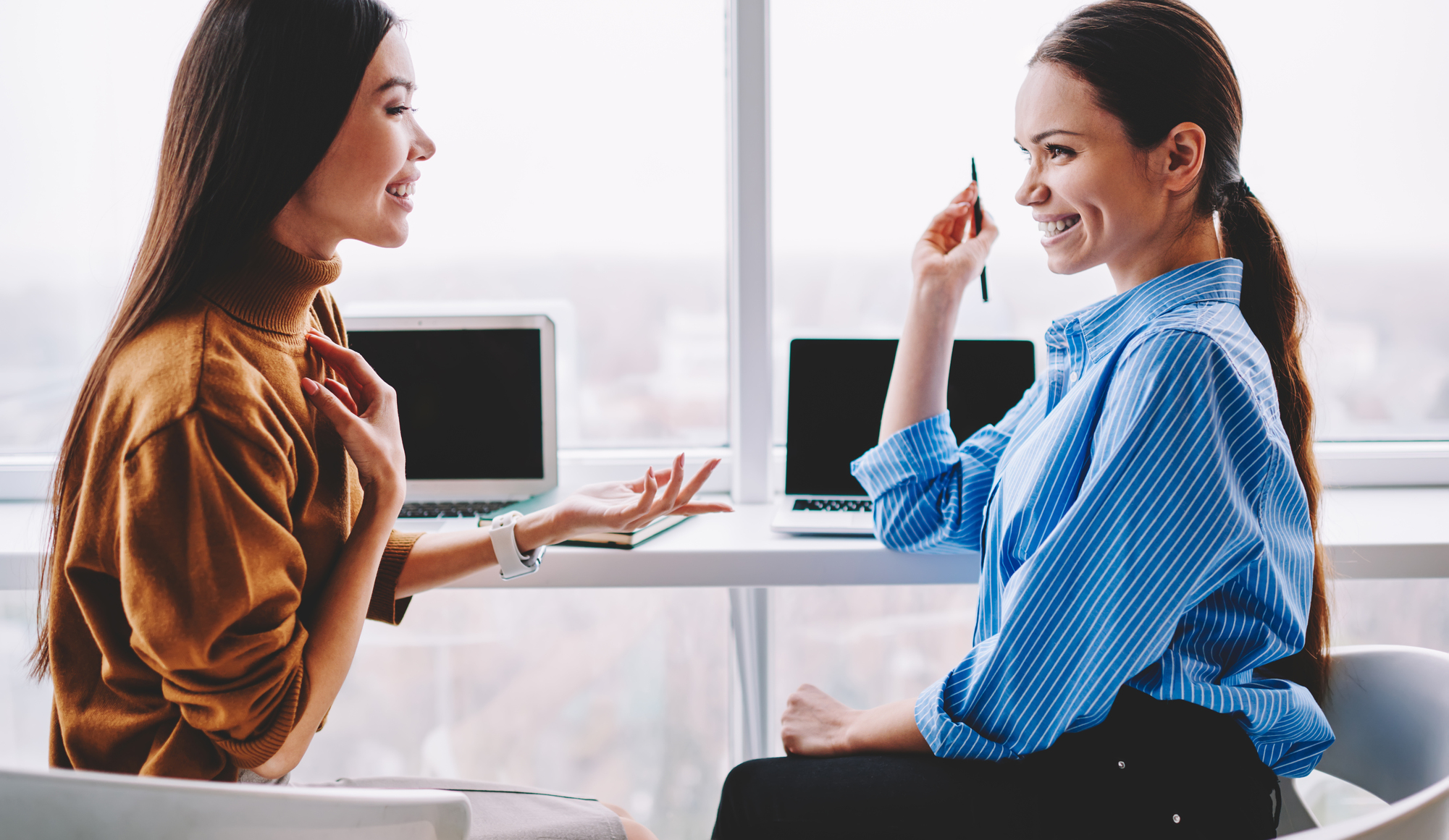 Two women are sitting at a desk with laptops, engaged in a friendly conversation. One is wearing a brown sweater and the other a blue striped shirt. They are in a bright office with a large window in the background.