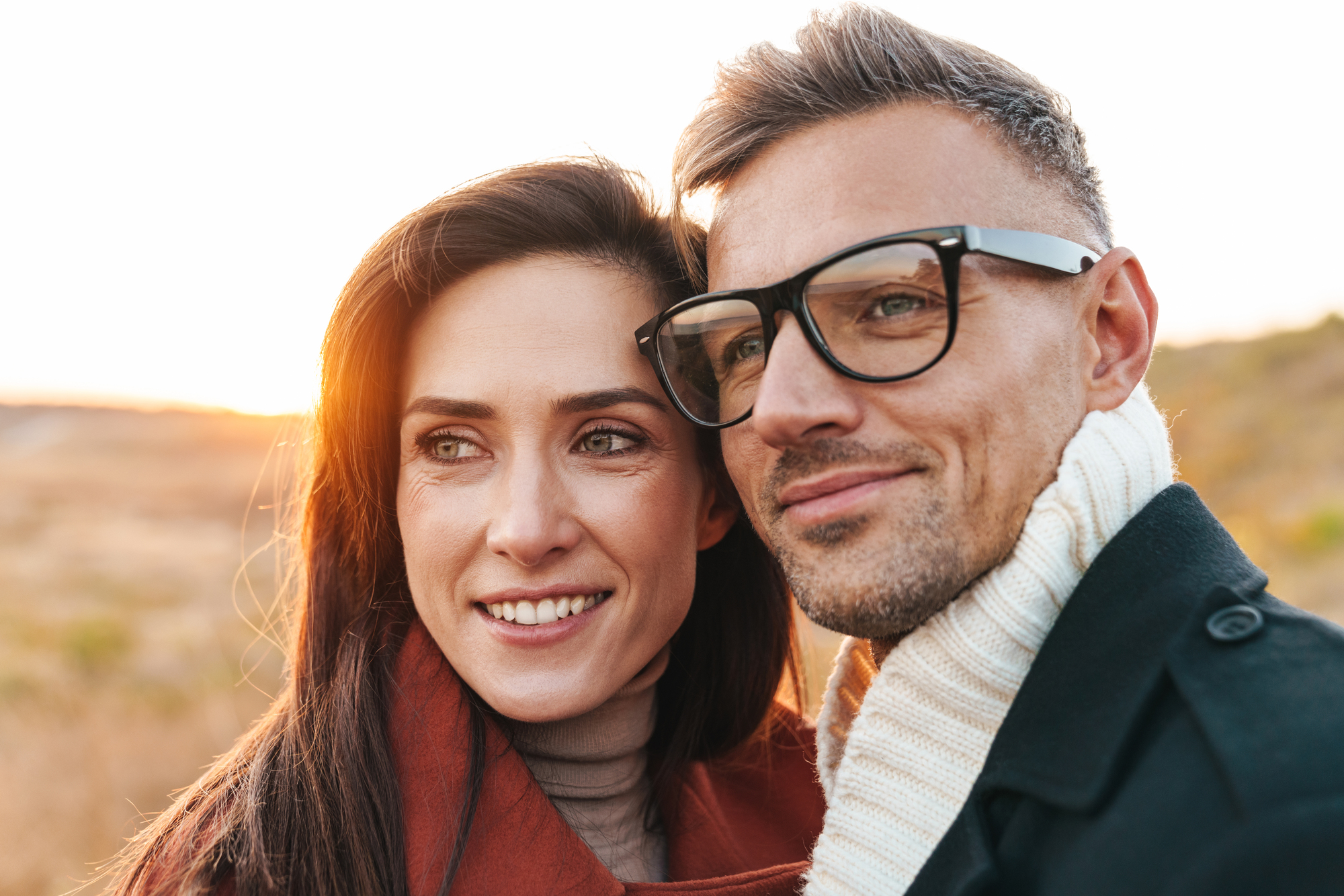 A couple smiling and looking into the distance, standing outdoors in a natural setting. The woman is wearing a rust-colored coat, and the man is wearing glasses and a scarf. The sun is setting in the background, casting a warm glow.