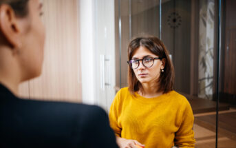 A woman wearing glasses and a yellow sweater listens attentively in an office setting. She stands near a glass partition while facing another person whose back is turned to the camera.