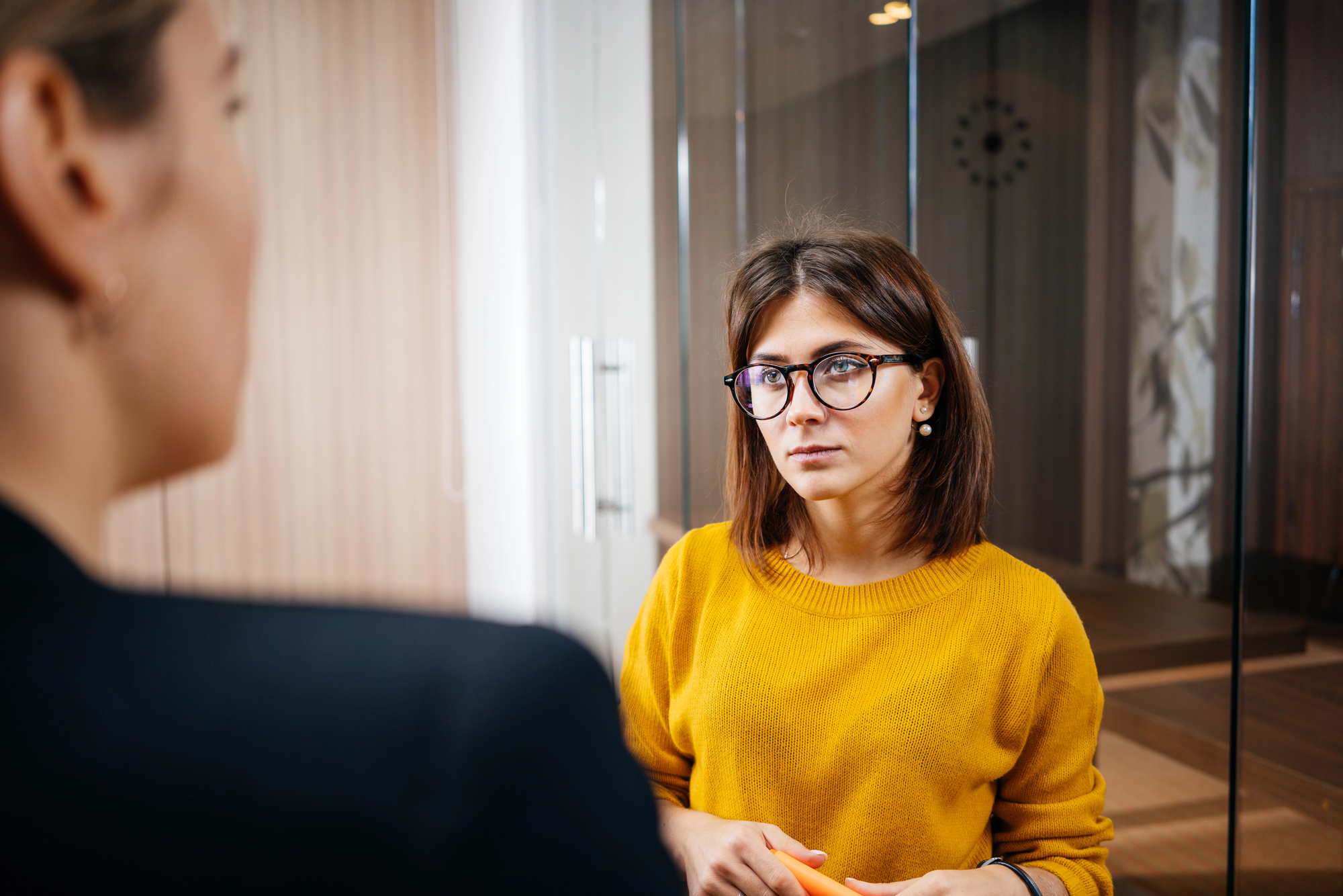 A woman wearing glasses and a yellow sweater listens attentively in an office setting. She stands near a glass partition while facing another person whose back is turned to the camera.