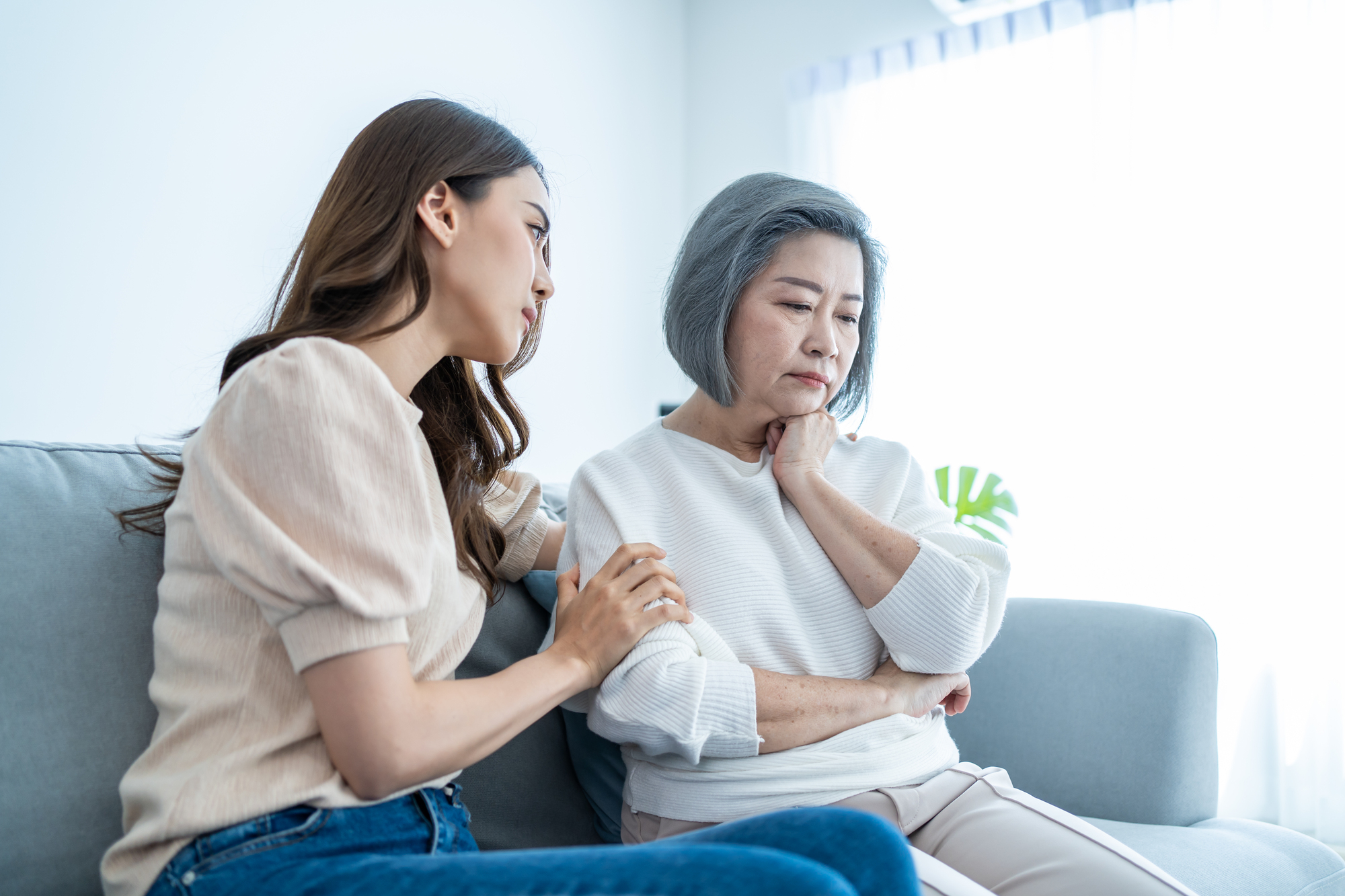 A young woman sits on a couch, gently comforting an older woman who appears upset. The older woman has gray hair and is wearing a white sweater, while the young woman has long brown hair and is wearing a light-colored shirt and jeans.