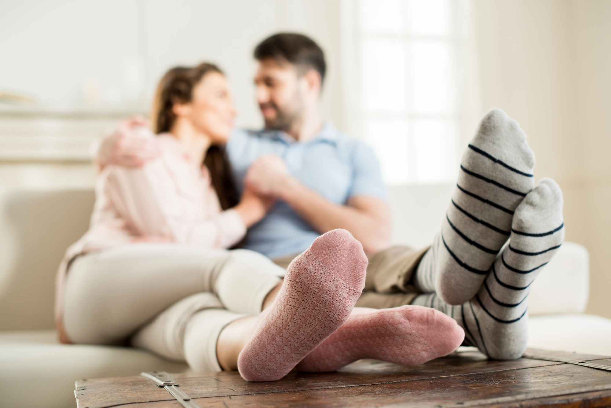 A couple is sitting on a couch, smiling and embracing each other. Their feet in cozy socks are in the foreground, with the focus on pink and striped patterns. The background is softly lit, creating a warm and relaxed atmosphere.