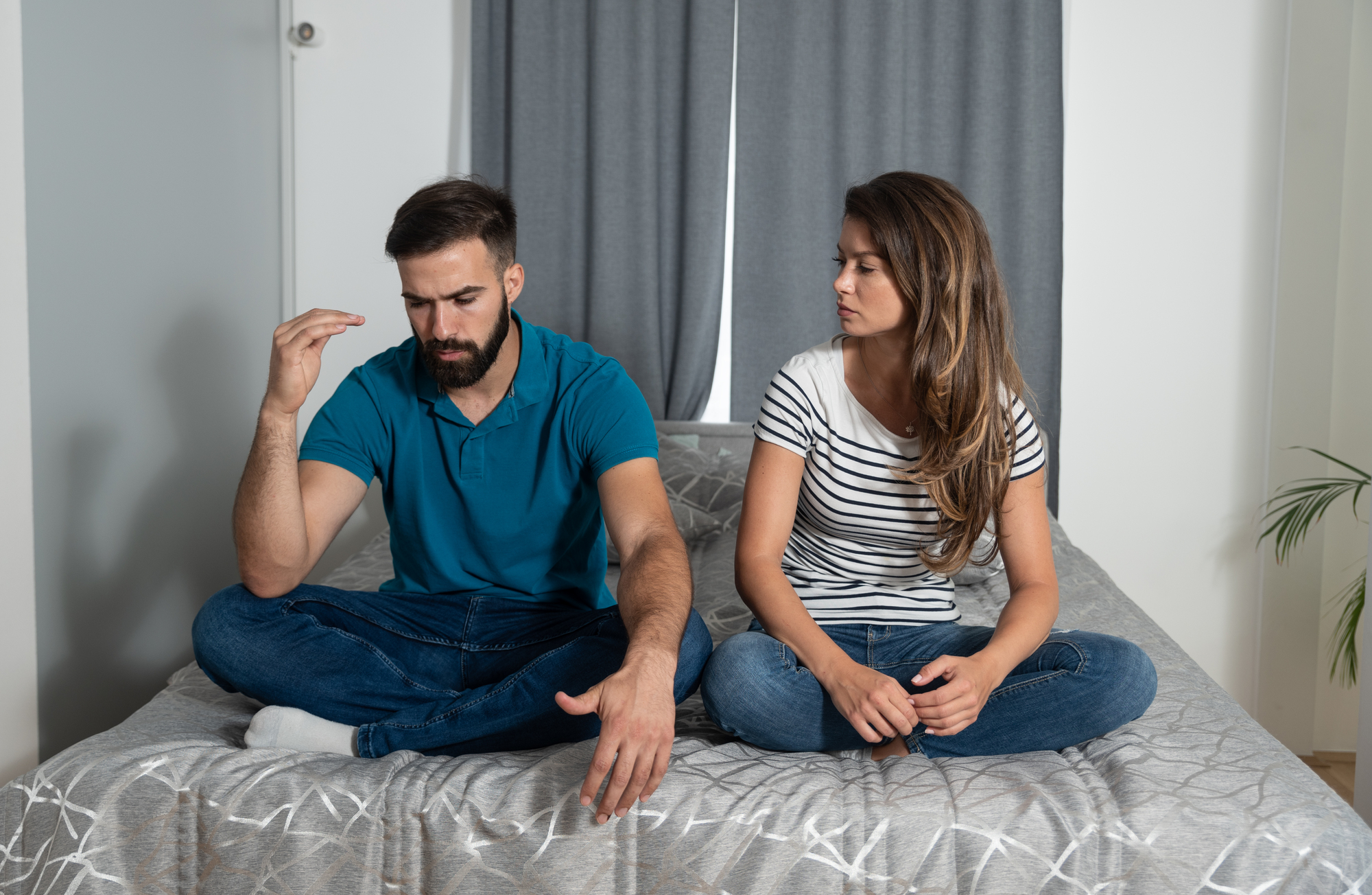 A man and woman sit cross-legged on a bed in a serious conversation. The man gestures with his hand while the woman listens attentively. Both wear casual clothing, and a plant is visible in the background.