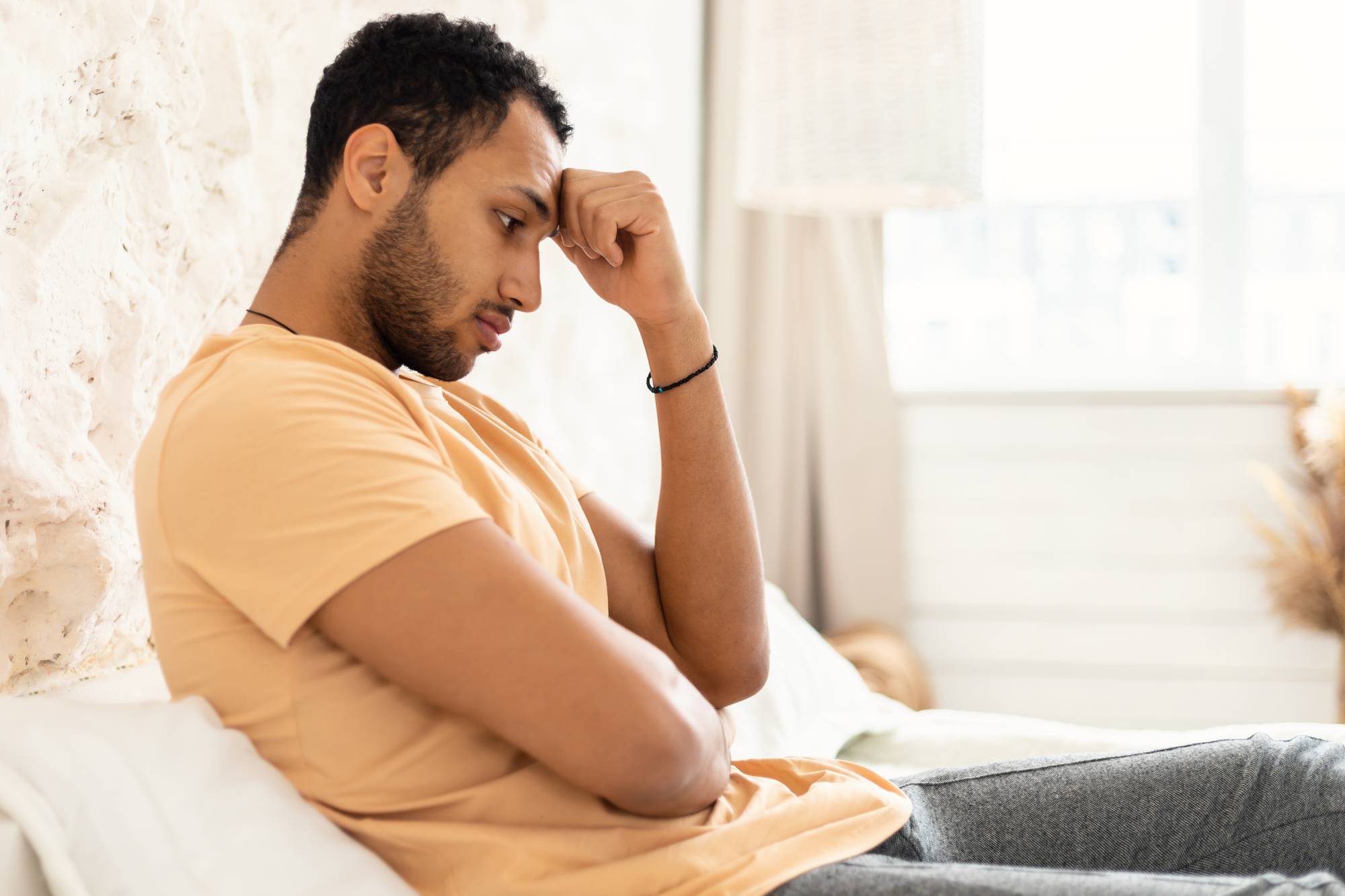 A person sits on a couch with their head resting on one hand, appearing deep in thought. They are wearing a light brown t-shirt and gray pants, with a light background and soft natural lighting.